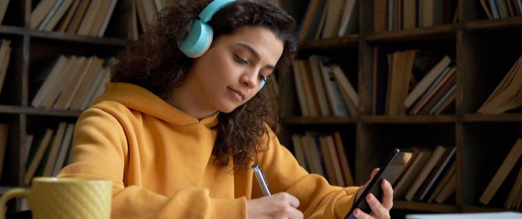 Teenage girl in yellow sweatshirt and blue headphones is holding her phone and writing things down.